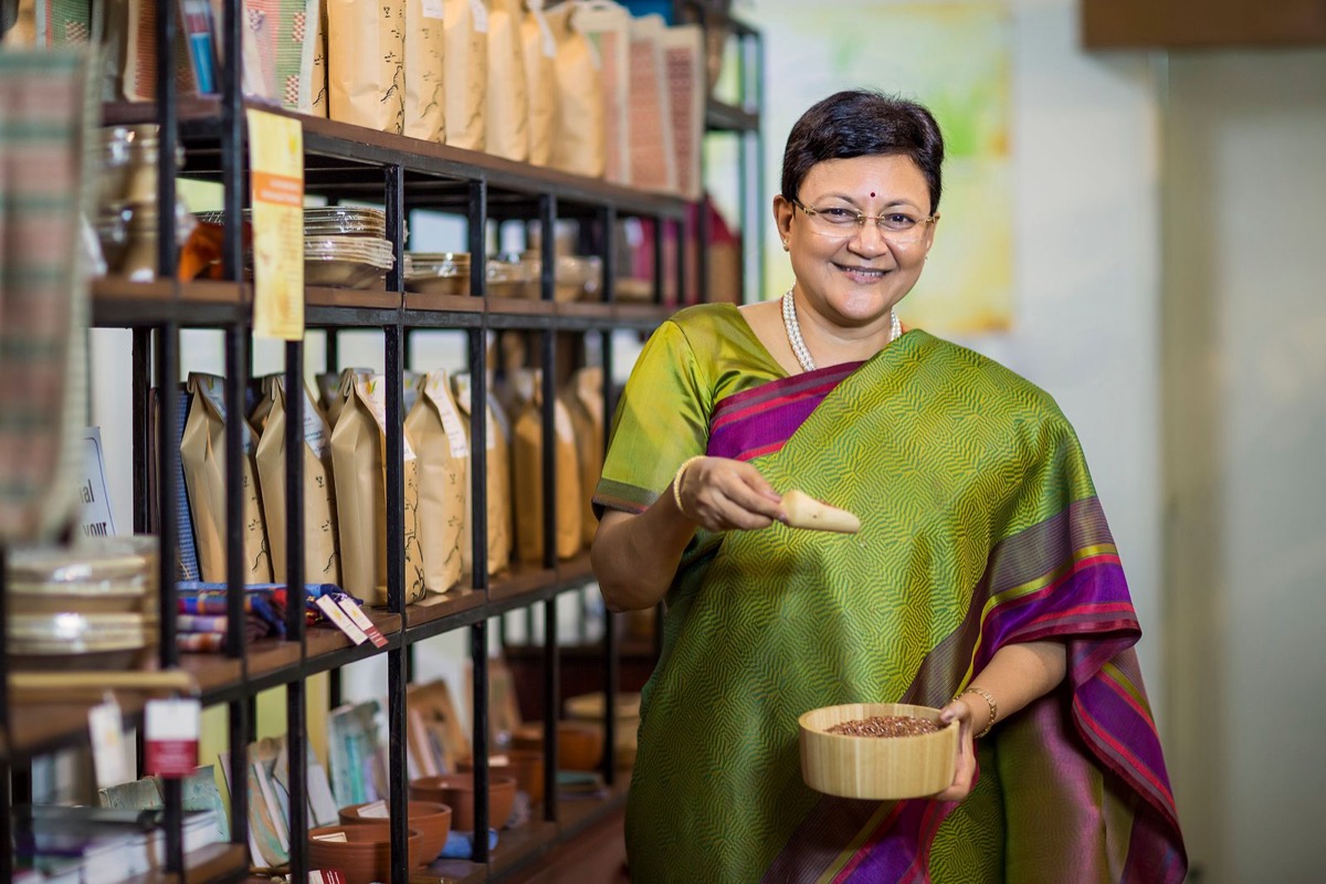 Smiling shop owner in a green silk saree holding a scoop of spices in her store